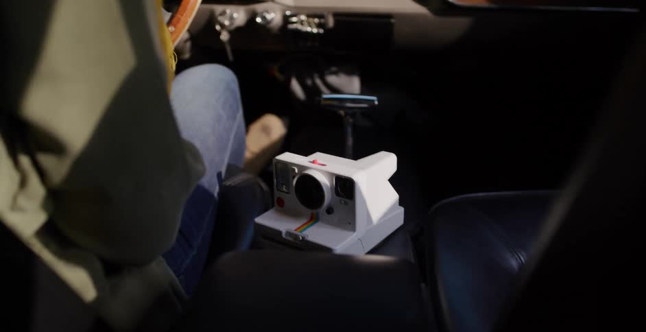 A vintage instant camera sits on the center console of a classic car during a sunny road trip