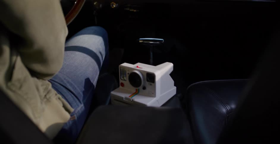 A person drives a vintage car through a sun-dappled forest, with a Polaroid camera on the console
