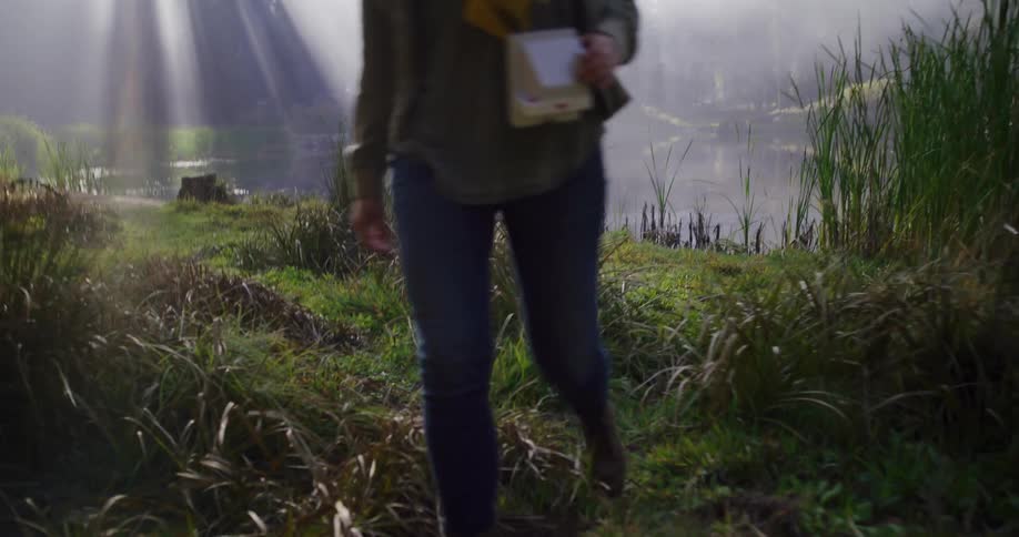 A person in hiking boots walks along a misty lakeside path, carrying a vintage portable computer as sun rays break through the fog