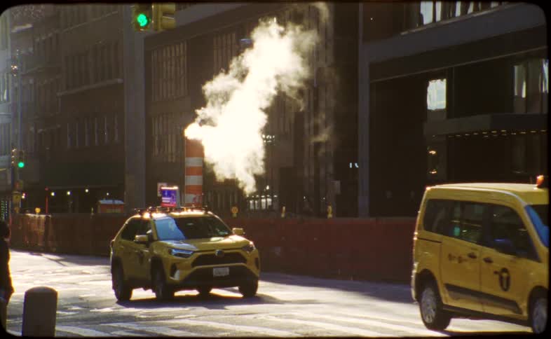 Iconic yellow cabs drive in slow motion past a steaming vent pipe on a sunlit street in New York City