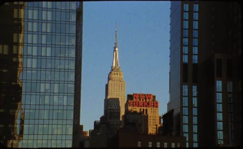 A vintage film shot tilts down revealing the Empire State Building and the iconic New Yorker Hotel sign against a clear blue sky
