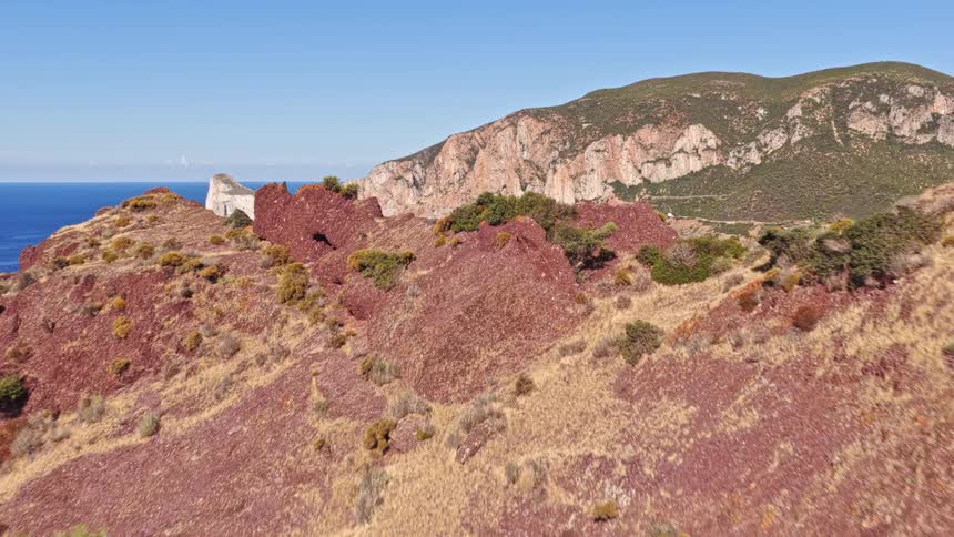 An aerial shot reveals a beautiful blue bay and the Pan di Zucchero sea stack from behind a red rocky hillside on the Sardinian coast