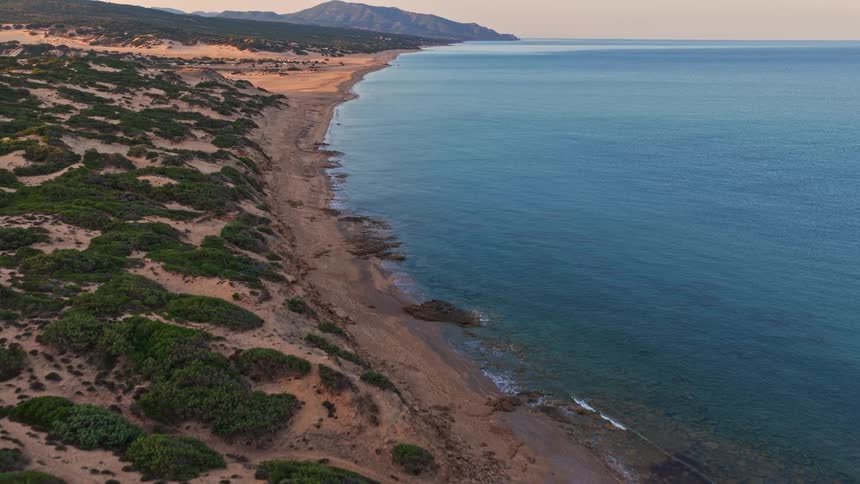 An aerial drone shot flies over a lush, green coastal landscape, revealing a long, secluded sandy beach and calm turquoise sea at sunrise