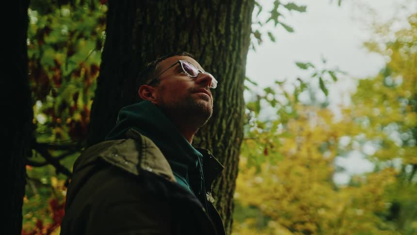 A thoughtful man wearing glasses finds a moment of peace while leaning against a tree in a colorful autumn forest