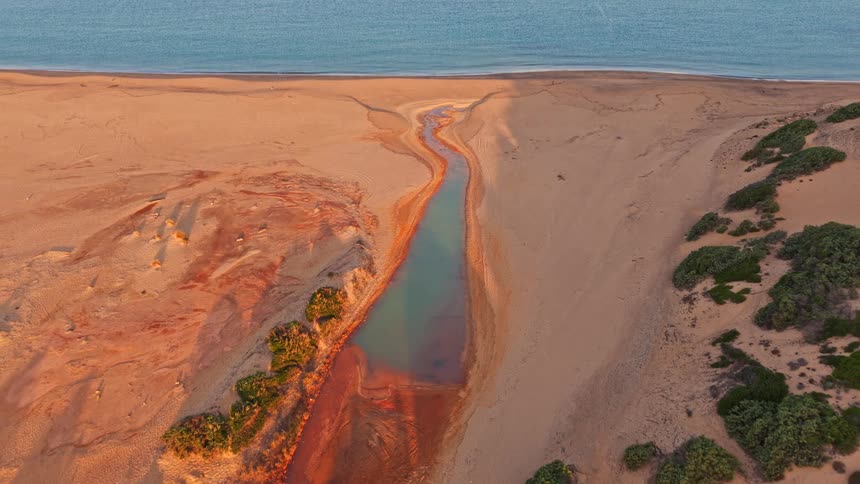 An aerial view pulls back to reveal a colorful river winding through a vast sandy desert landscape and emptying into the ocean at sunset