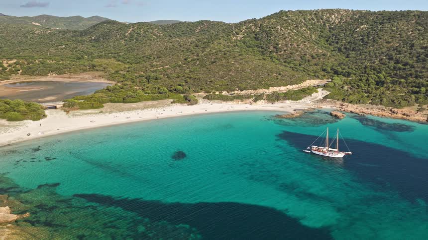 Stunning aerial view rises above a secluded turquoise cove where a classic sailboat is anchored off a pristine white sand beach