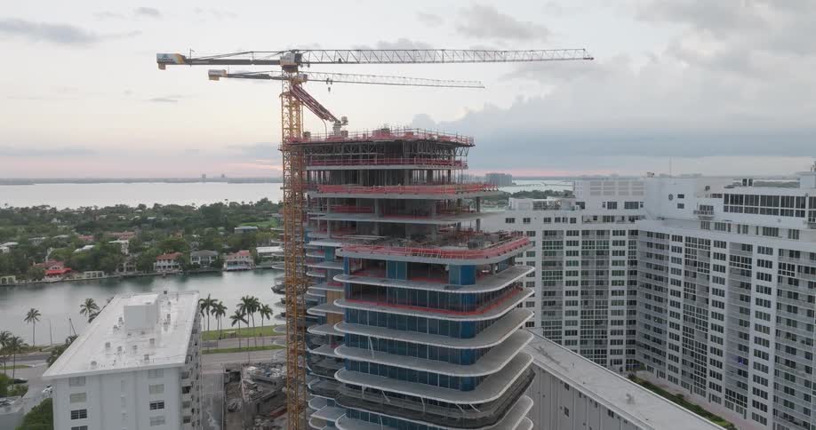 A slow-moving aerial shot reveals a modern high-rise building under construction with cranes at dusk, surrounded by a coastal cityscape