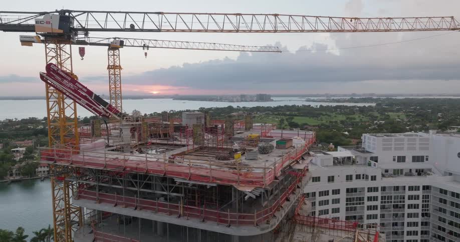 Aerial footage pulls back from two large construction cranes to reveal a high-rise building site overlooking a coastal city at sunset
