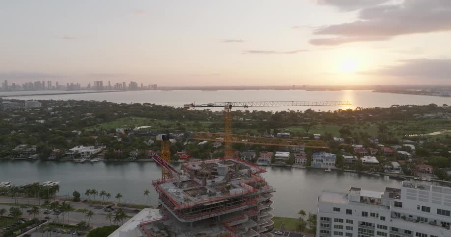 Aerial pullback reveals a high-rise building under construction with cranes overlooking the water with the city skyline in the distance at sunrise