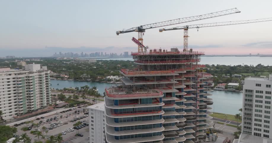 An aerial drone circles a high-rise building under construction with two large cranes overlooking the ocean and city at sunset