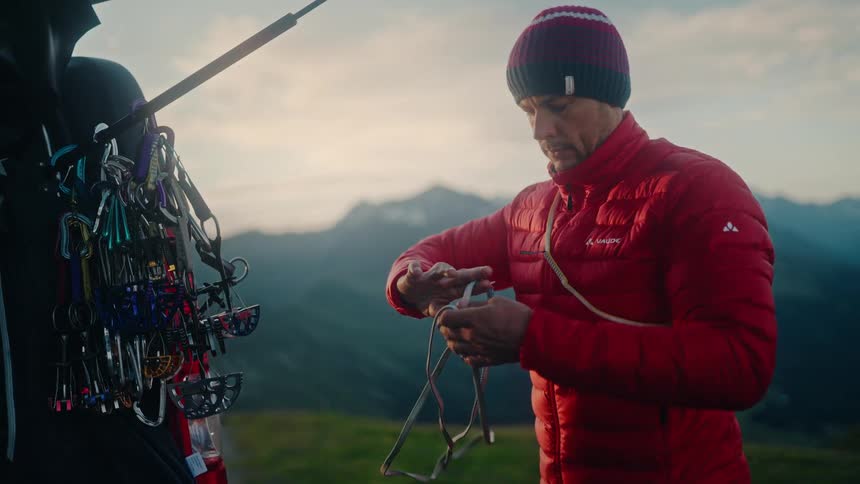 A male mountaineer in a red jacket prepares his climbing rope and equipment from his vehicle with a stunning mountain view at dawn