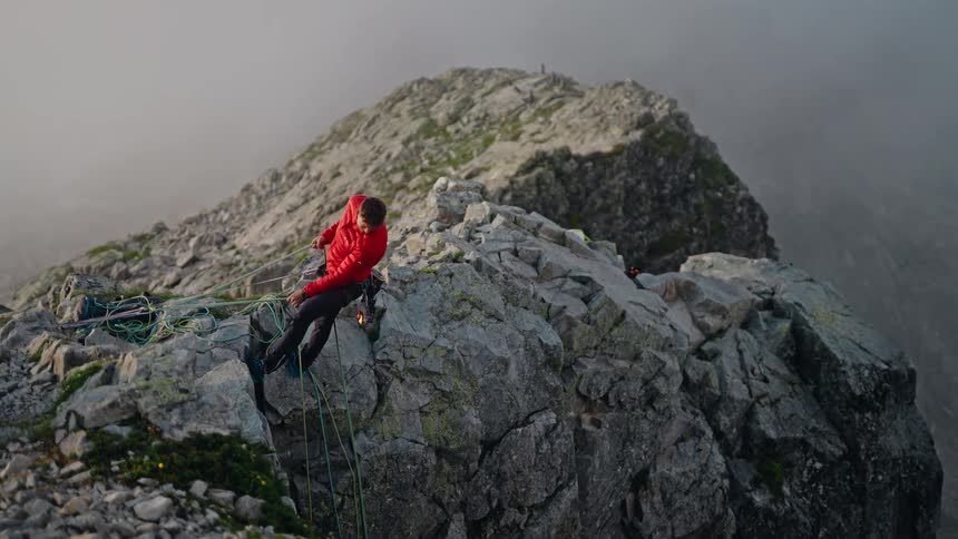 A male climber in a red jacket prepares his ropes while sitting on a steep, rocky mountain ridge surrounded by thick fog