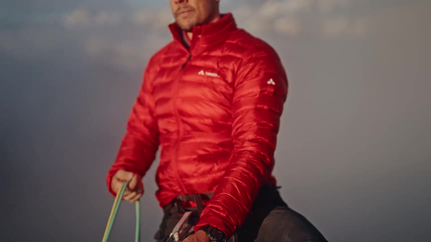 An alpinist in a red jacket holds his climbing ropes while standing on a mountain summit above the clouds at golden hour