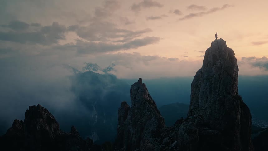 A silhouetted hiker with a backpack triumphantly stands atop a rugged mountain peak at sunset, overlooking a vast, cloud-filled valley