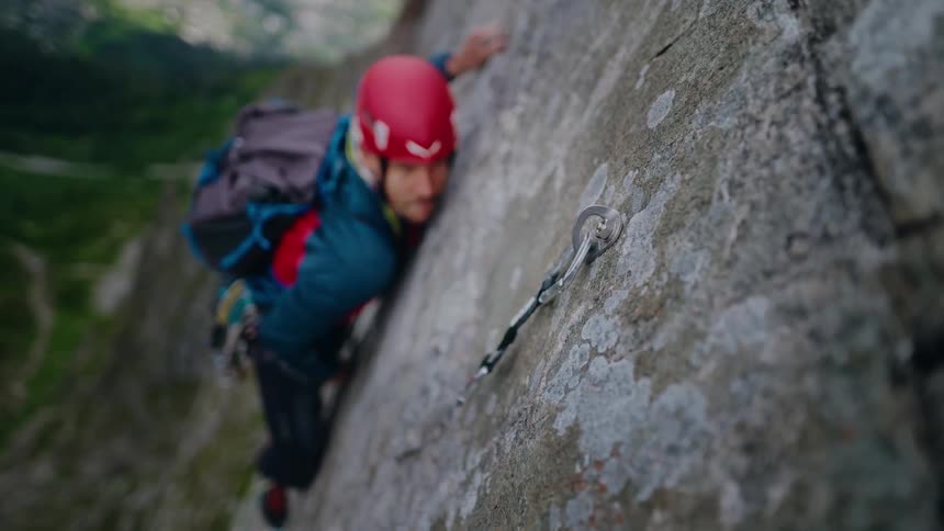 A determined male rock climber in a red helmet carefully ascends a sheer mountain face, clipping his safety rope into an anchor