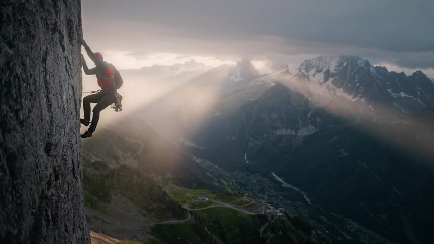 A lone rock climber scales a sheer cliff face as dramatic sun rays illuminate an epic mountain range and valley below