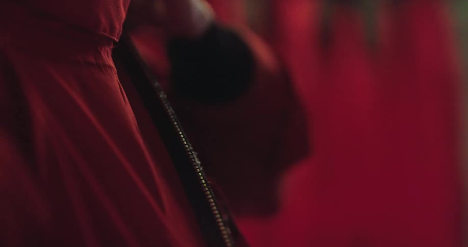 A close-up slow motion shot shows a person zipping up their red jumpsuit in a dimly lit, moody environment
