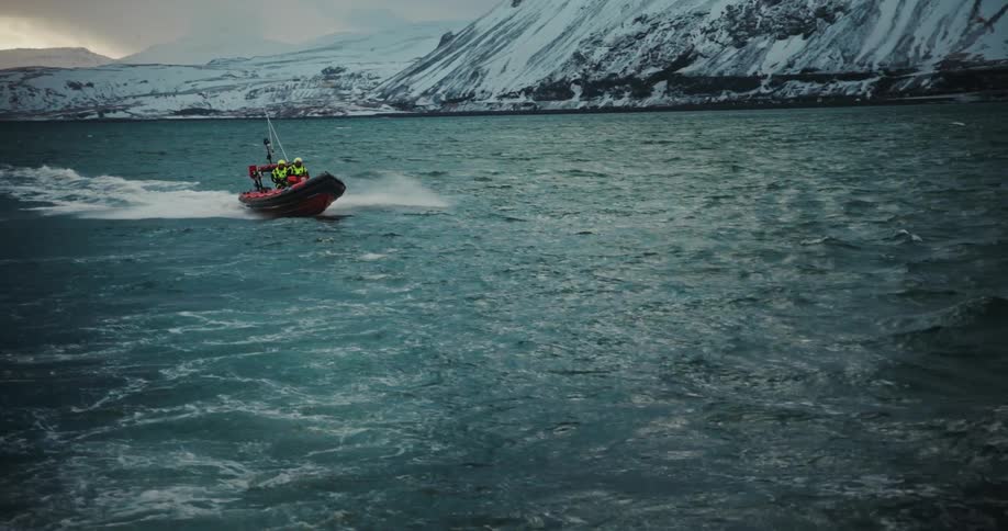 A search and rescue team speeds across the choppy arctic sea in a rigid inflatable boat against a dramatic backdrop of snow-covered mountains