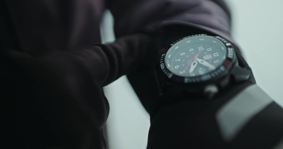 A person wearing a black glove and winter jacket pulls back their sleeve to check a tactical analog watch in a slow-motion close-up
