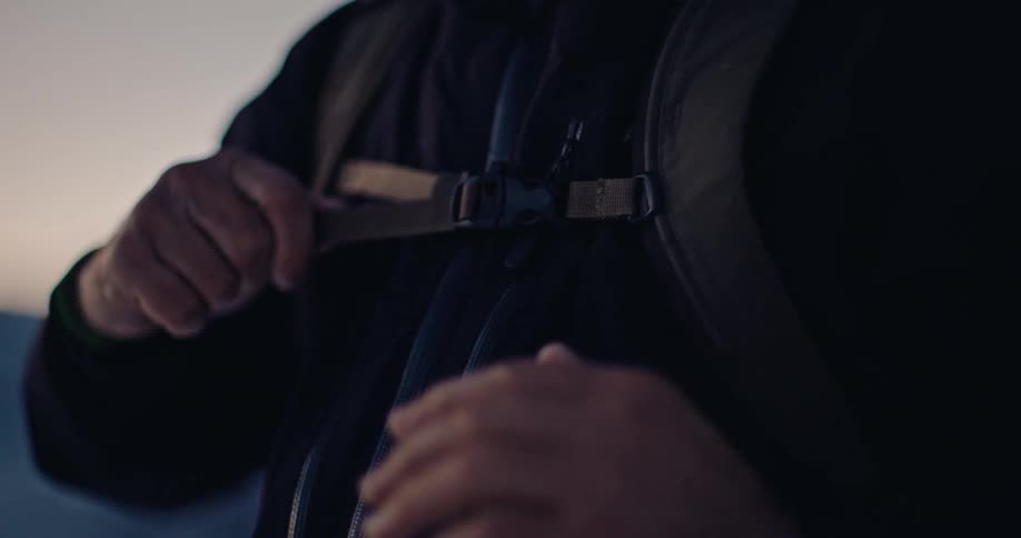 An adventurer carefully fastens the chest strap of a backpack in a moody slow-motion close-up