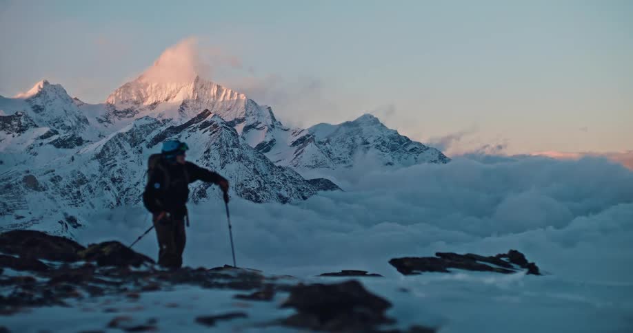 A ski mountaineer hikes along a snowy ridge above a sea of clouds as the sun sets on the majestic mountain peaks