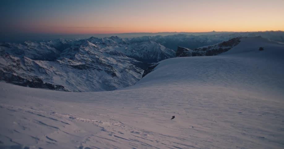 A lone skier carves fresh tracks down a snowy mountain slope in slow motion, revealing a breathtaking panorama of the Alps at sunset