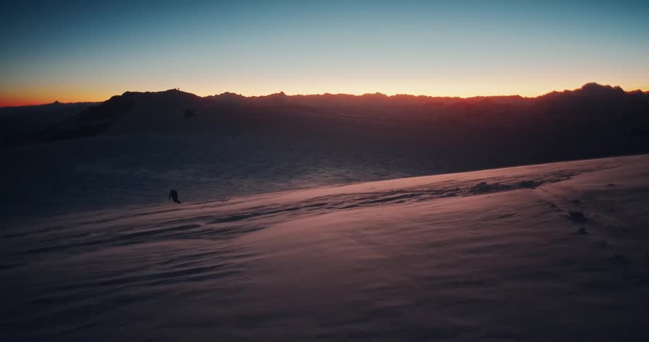 A silhouetted backcountry skier treks across a vast, windy, snow-covered mountain range at sunrise
