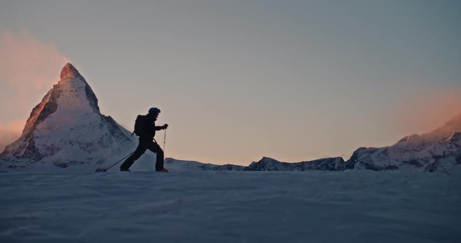 In slow motion, a silhouetted hiker treks across a snowy landscape with the iconic Matterhorn in the background at sunset
