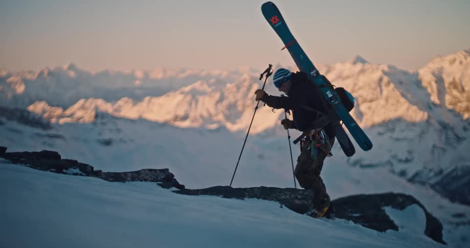 A determined backcountry skier hikes up a snowy mountain ridge in slow motion with a stunning alpine sunrise in the background