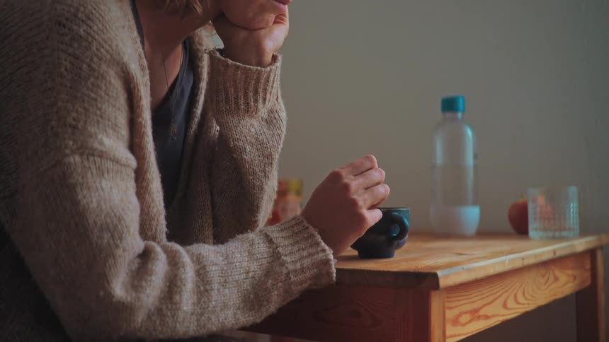 A contemplative woman in a cozy sweater sits alone at a wooden table, slowly stirring a drink in a mug