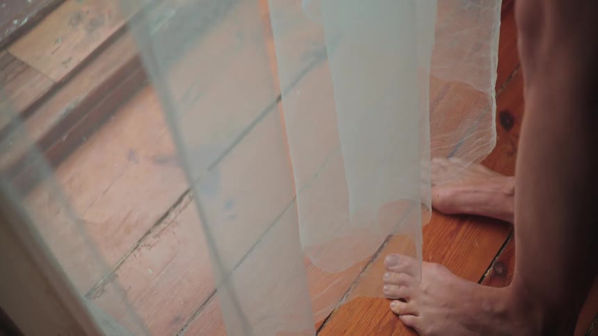 Bare feet stand on a rustic wooden floor next to a gently blowing sheer curtain in slow motion