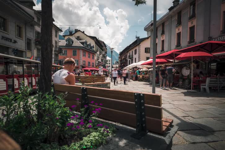 A slow dolly-out timelapse captures a bustling European town square with blurred pedestrians rushing past a man sitting peacefully on a bench