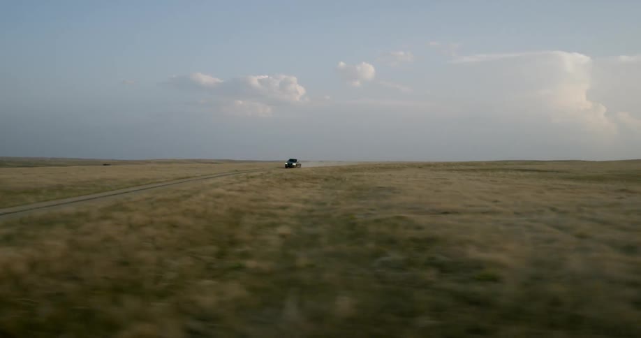 Panning quickly across and open field as a truck drives on a dusty dirt road