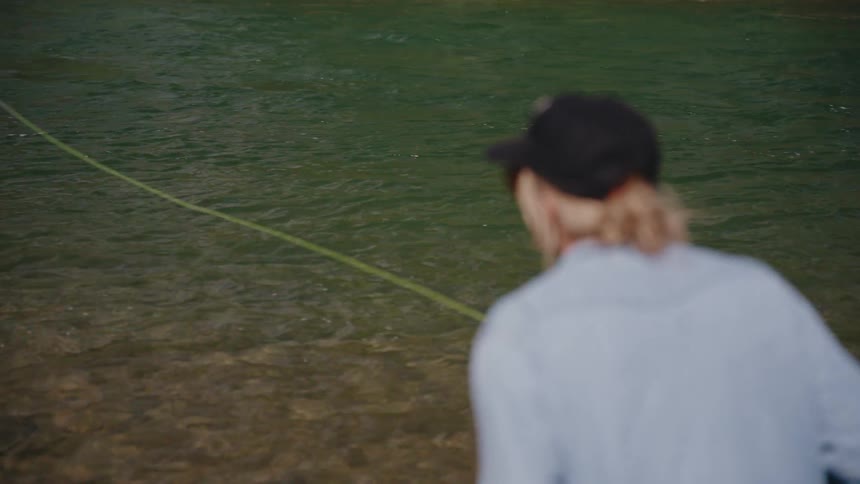Over the shoulder view of a fly fisherman casting their line into a flowing river in slow motion