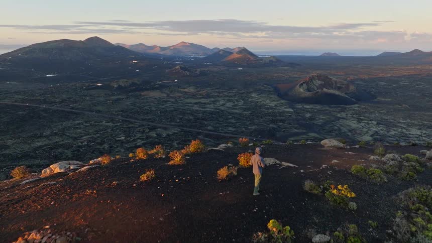 An aerial view shows a lone hiker walking along the summit of a volcano, revealing the vast and beautiful volcanic landscape of Lanzarote at sunrise