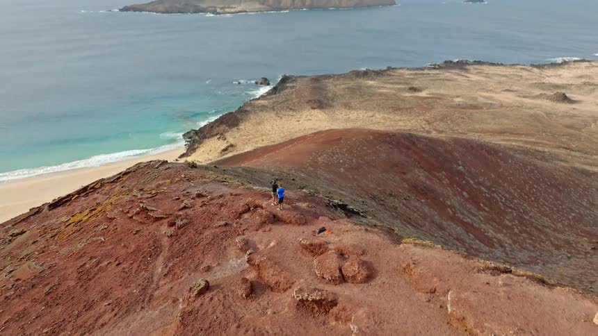 Aerial shot reveals two hikers on the summit of a volcanic mountain overlooking a beautiful sandy beach and a remote island
