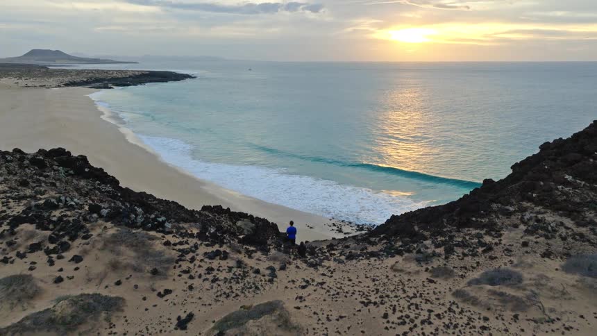 An aerial shot tilts up from a remote campsite on a sandy cliff to reveal a stunning sunset over a pristine beach and ocean