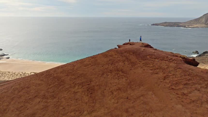 An aerial drone flies over a runner and his dog as they ascend a red volcanic cone, revealing a stunning secluded beach with turquoise water