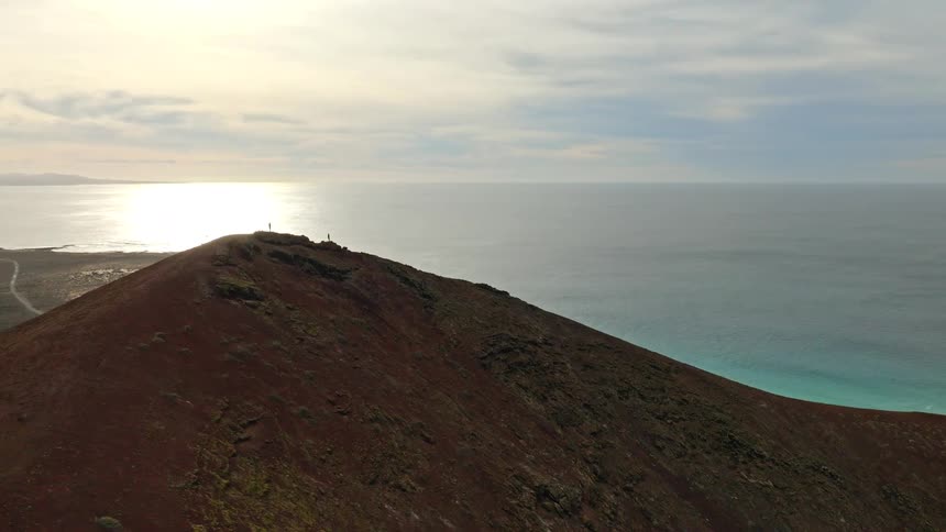 An aerial view reveals people standing on the summit of a volcanic mountain, gazing at the sunlit ocean and distant islands