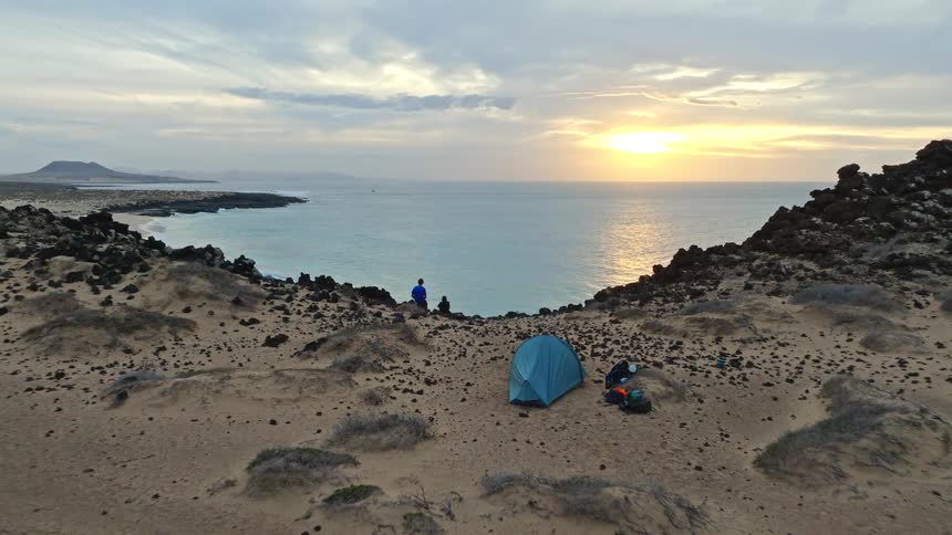 Aerial shot pulls back to reveal a couple camping on a cliff, watching the sunset over a beautiful, secluded beach