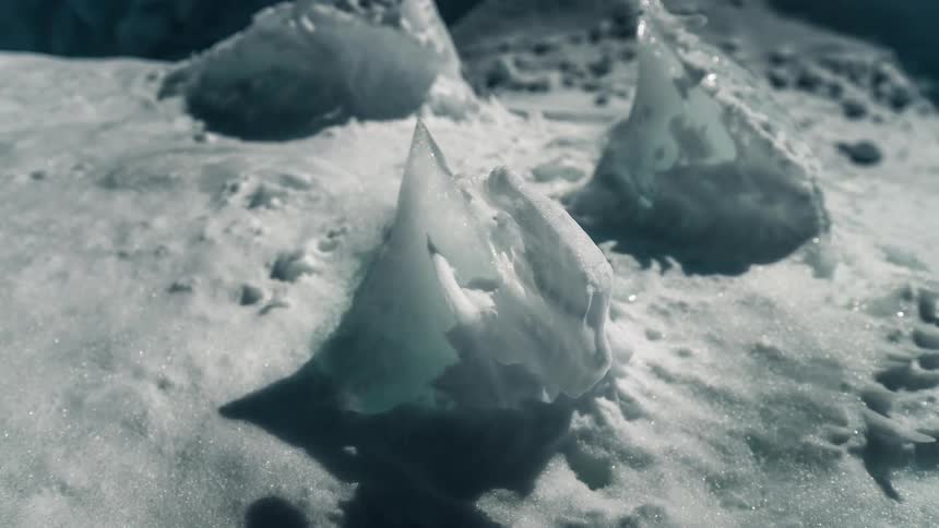 A close-up time-lapse shows a sharp, crystalline ice shard slowly melting in the bright sunlight on a frozen landscape