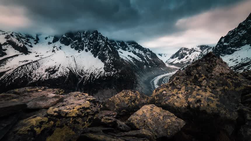 A dramatic time-lapse of dark, moody clouds parting to reveal a vast glacier winding its way through a rugged, snow-capped mountain range
