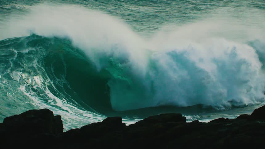 A huge turquoise ocean wave crests and breaks in dramatic slow motion behind silhouetted rocks
