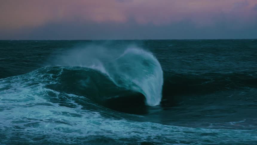 A massive, powerful ocean wave crashes in super slow motion under a dramatic, moody sky