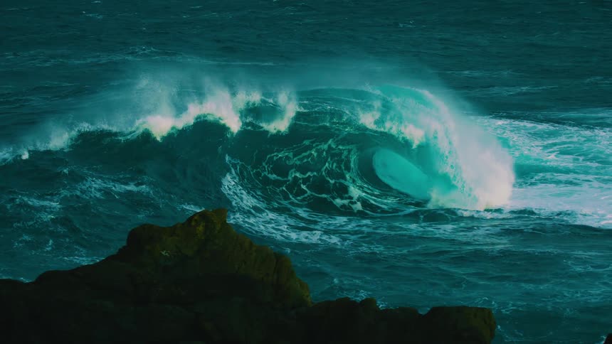 Slow motion footage captures the immense power of a giant, glowing turquoise wave breaking in a dark sea