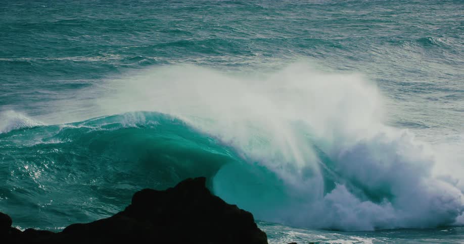 A giant turquoise ocean wave crashes in slow motion behind silhouetted rocks