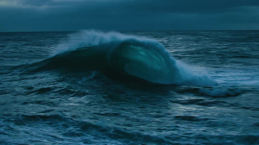 A powerful ocean wave builds and crashes in dramatic slow motion under a dark, moody sky