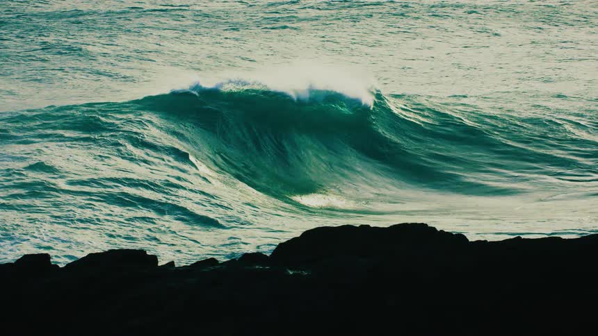 An enormous teal-colored ocean wave builds and crashes in dramatic super slow motion behind silhouetted rocks
