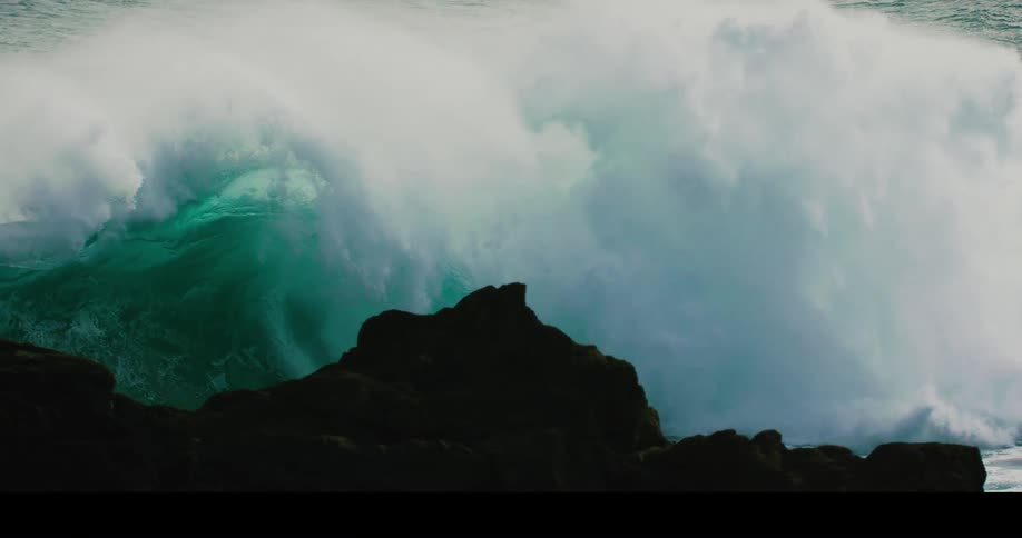 A massive turquoise ocean wave curls and breaks in dramatic slow motion behind silhouetted coastal rocks