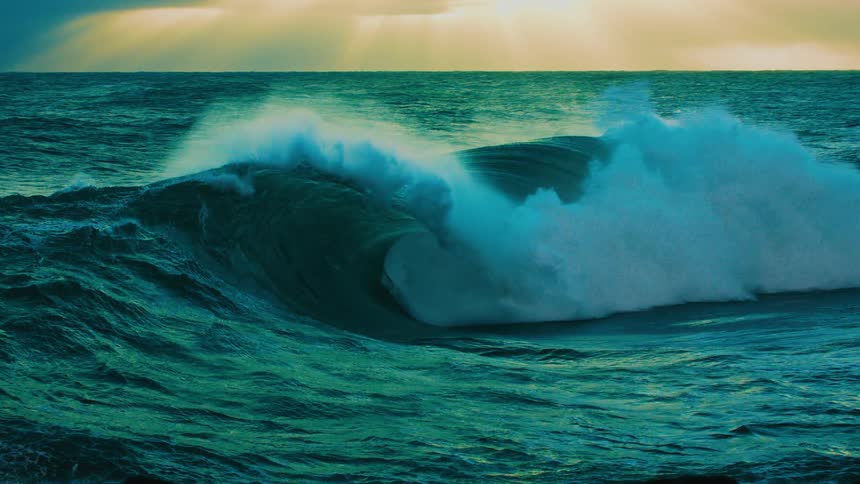 A powerful turquoise ocean wave curls and crashes in cinematic slow motion under a dramatic, glowing sky